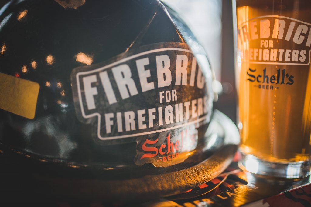Firefighter helmet, with a Schell's Firebrick for Firefighters sticker, next to a branded Firebrick for Firefighters pint glass full of beer